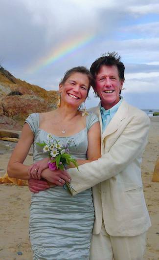 John Moore and Andrea Geyling-Moore holding each other on their Martha's Vineyard wedding day in 2013 with a sliver of rainbow behind them peeking over the beach cliff.