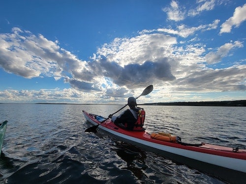 John Moore Jr kayaking on Chilmark Pond from behind, with expansive blue sky and fluffy clouds going into the horizon.