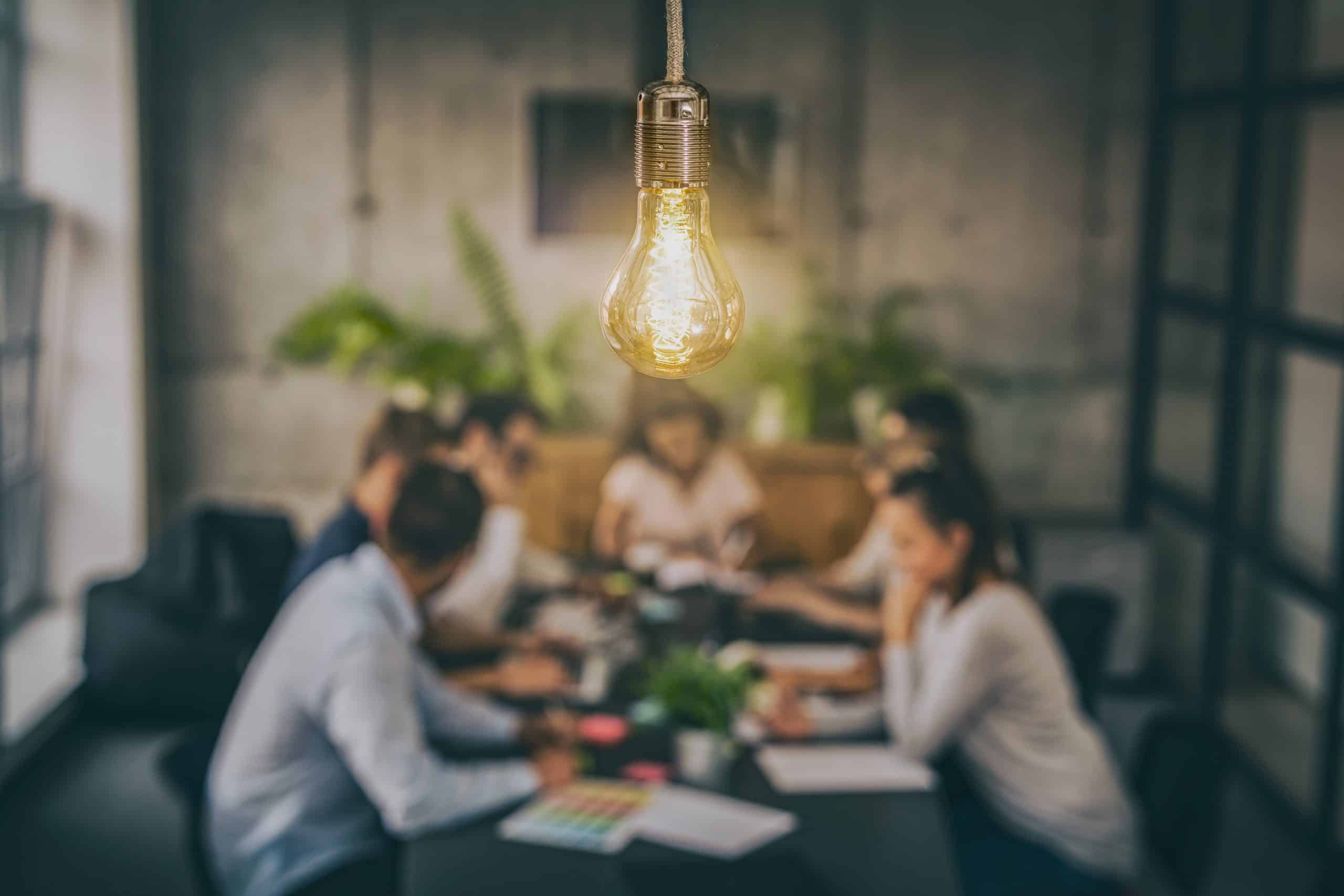 Group of young business people out of focus in the background look excited while meeting together. A glowing light bulb as a new idea is in focus in the foreground.